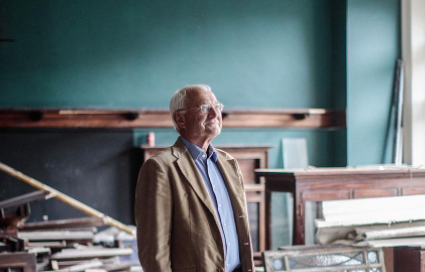A man with white hair and glasses stands in front of a blue wall, with wooden planks, covered up furniture and other evidence of construction in progress in the background