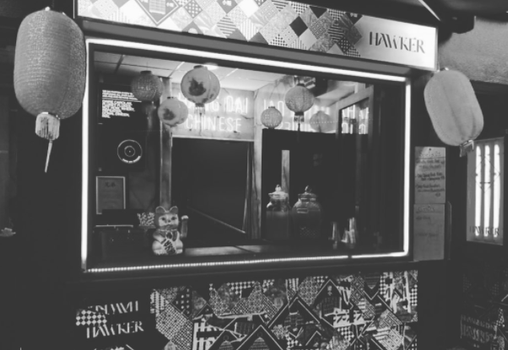 Black and white image of Hawker, a takeaway hatch with chinese lanterns, jars of food on the counter and a neon "Hang Dai" sign in the background