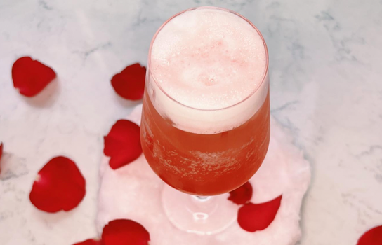 pink cocktail in a champagne glass with foam on top, with rose petals scattered in the background