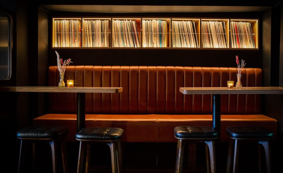 tan leather bar couch with high tables and stools in front of it, shelves stacked with vinyl records in the background
