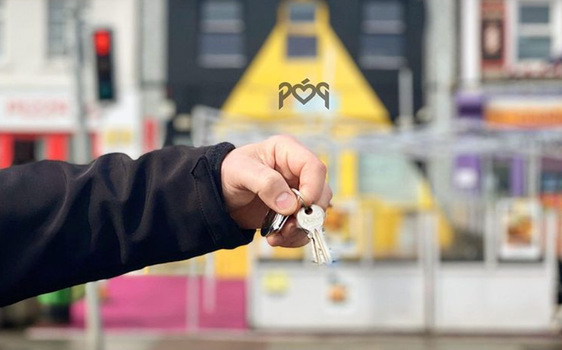 hand holding a set of keys in front of a row of shops with outdoor seating - the Póg logo can be seen in the background