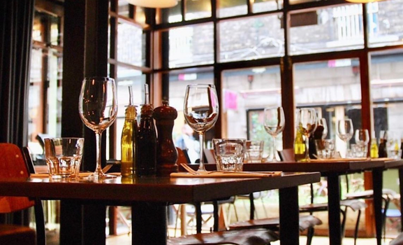 interior of a restaurant with large windows looking out to the street outside, with wine glasses, condiments and table settings