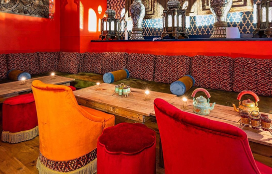 interior of electric ballroom with red and orange velvet seats and stools, a long wooden table with teapots, candles and glasses