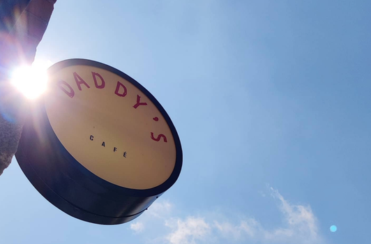 "Daddy's Cafe" round sign hanging from a red brick wall against a blue sky and the sun shining in the background