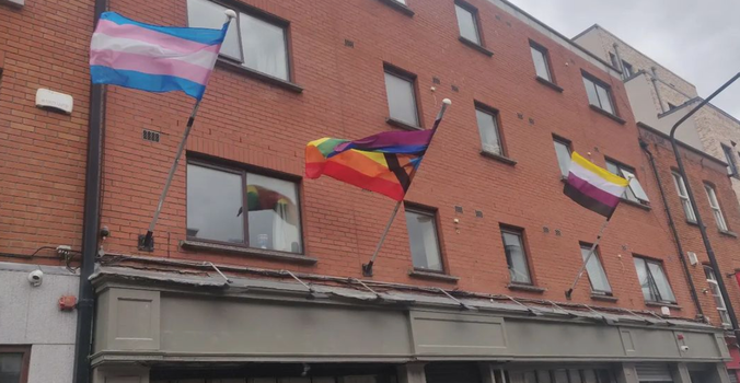 Trans flag and LGBTQ pride flag waving outside a shop front with red brick building above