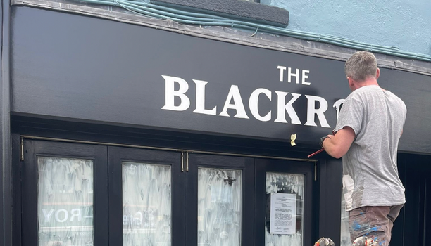 person painting signage which reads "The Blackrock" onto a black shop front