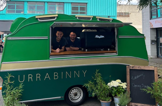 green and gold food caravan with "Currabinny" written on the side, two men standing at the hatch smiling for the camera