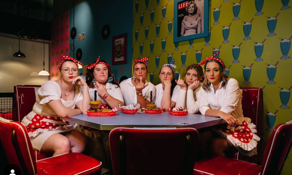 servers st Karen's diner sitting around a traditional diner-style table, wearing red and white polka dot aprons and headbands and looking disgruntled